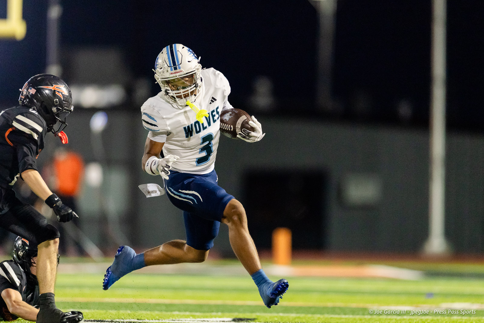 west-plains-vs-dumas-football-jpegJoe-pps-24 | Press Pass Sports Football player in a white and blue Wolves jersey with number 3 runs with the ball during a night game, pursued by a defender in black.