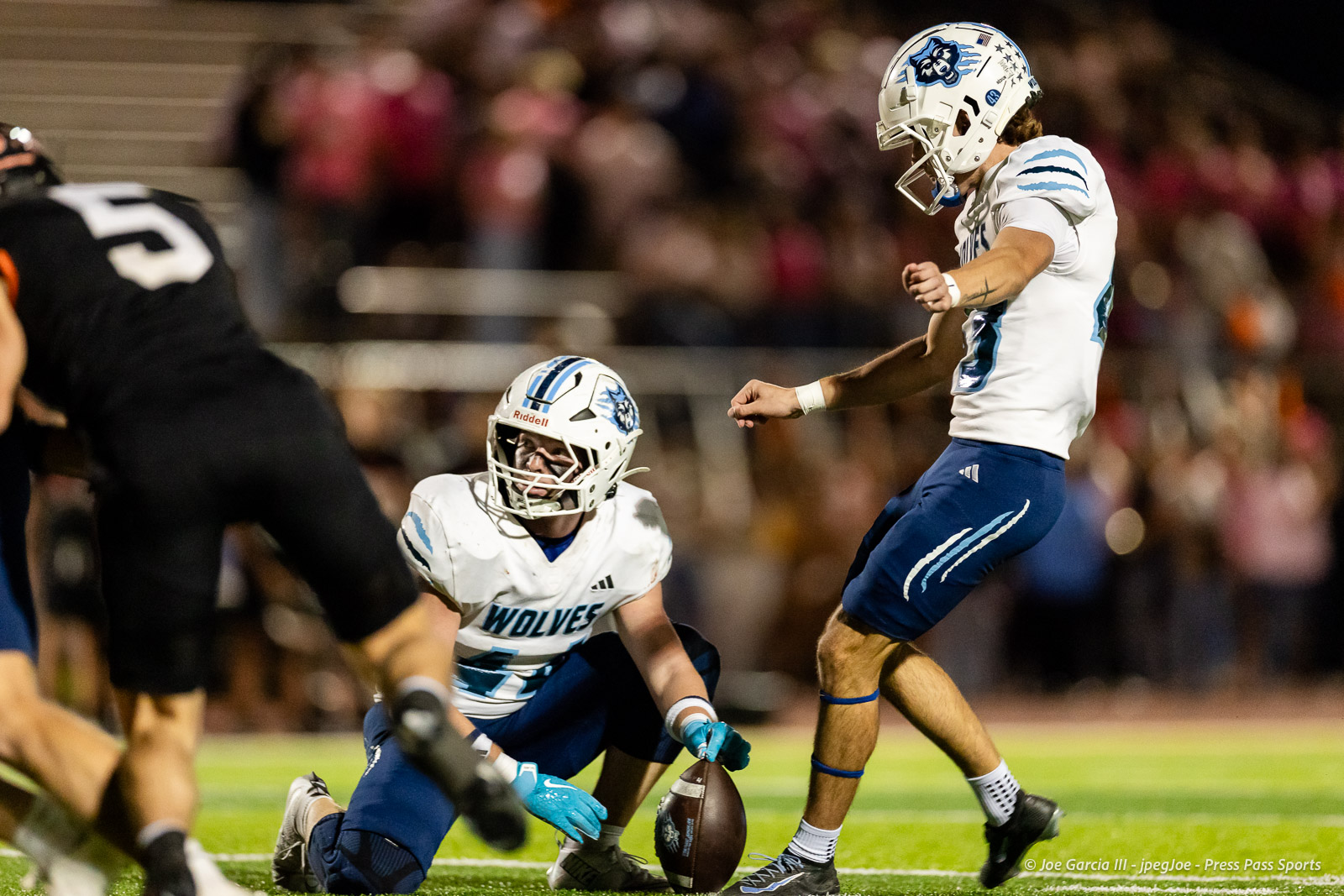 west-plains-vs-dumas-football-jpegJoe-pps-31 | Press Pass Sports Two American football players in white-and-blue Wolves uniforms compete for a loose ball on a night game, one on the ground reaching for the ball as a teammate approaches.