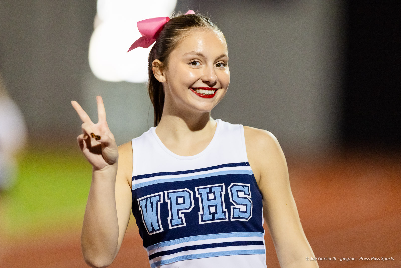 west-plains-vs-dumas-football-jpegJoe-pps-32 | Press Pass Sports Smiling cheerleader in a WPHS uniform making a peace sign, pink bow in her hair on an outdoor track.