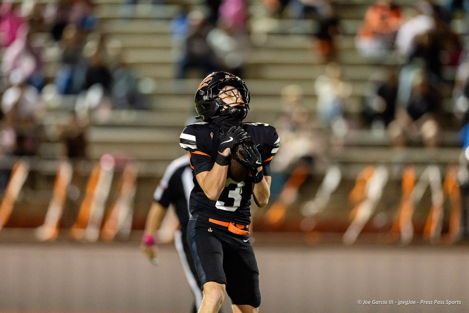west-plains-vs-dumas-football-jpegJoe-pps-34 | Press Pass Sports Football player in black uniform (#3) reaches for a catch on the field with a football near his chest; crowd and bleachers blurred in the background.