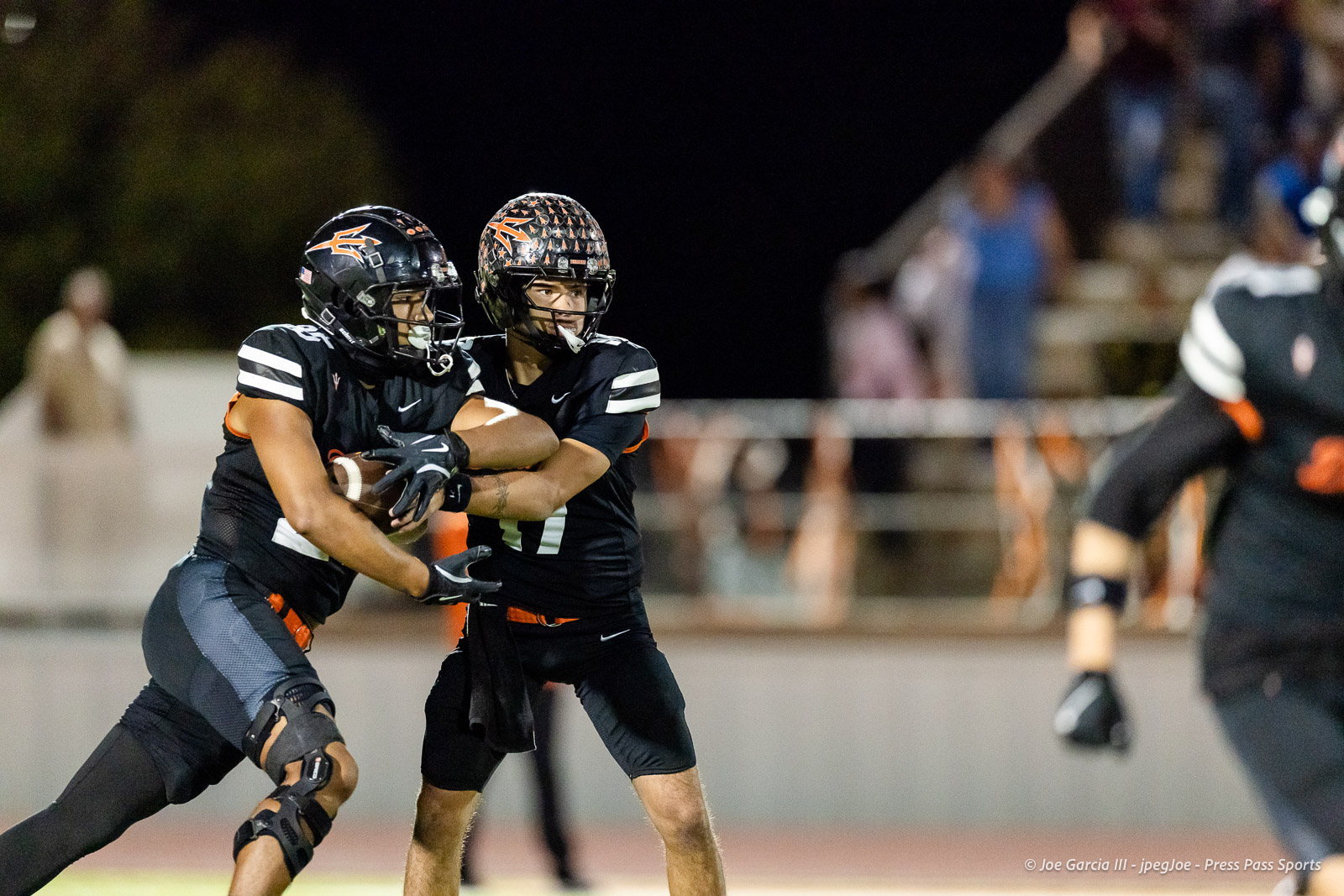 west-plains-vs-dumas-football-jpegJoe-pps-35 | Press Pass Sports Two football players in black uniforms tackle for the ball during a night game on a lit stadium field.