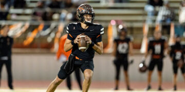 Quarterback in a black uniform runs with the football, looking downfield during a game on a lit field.