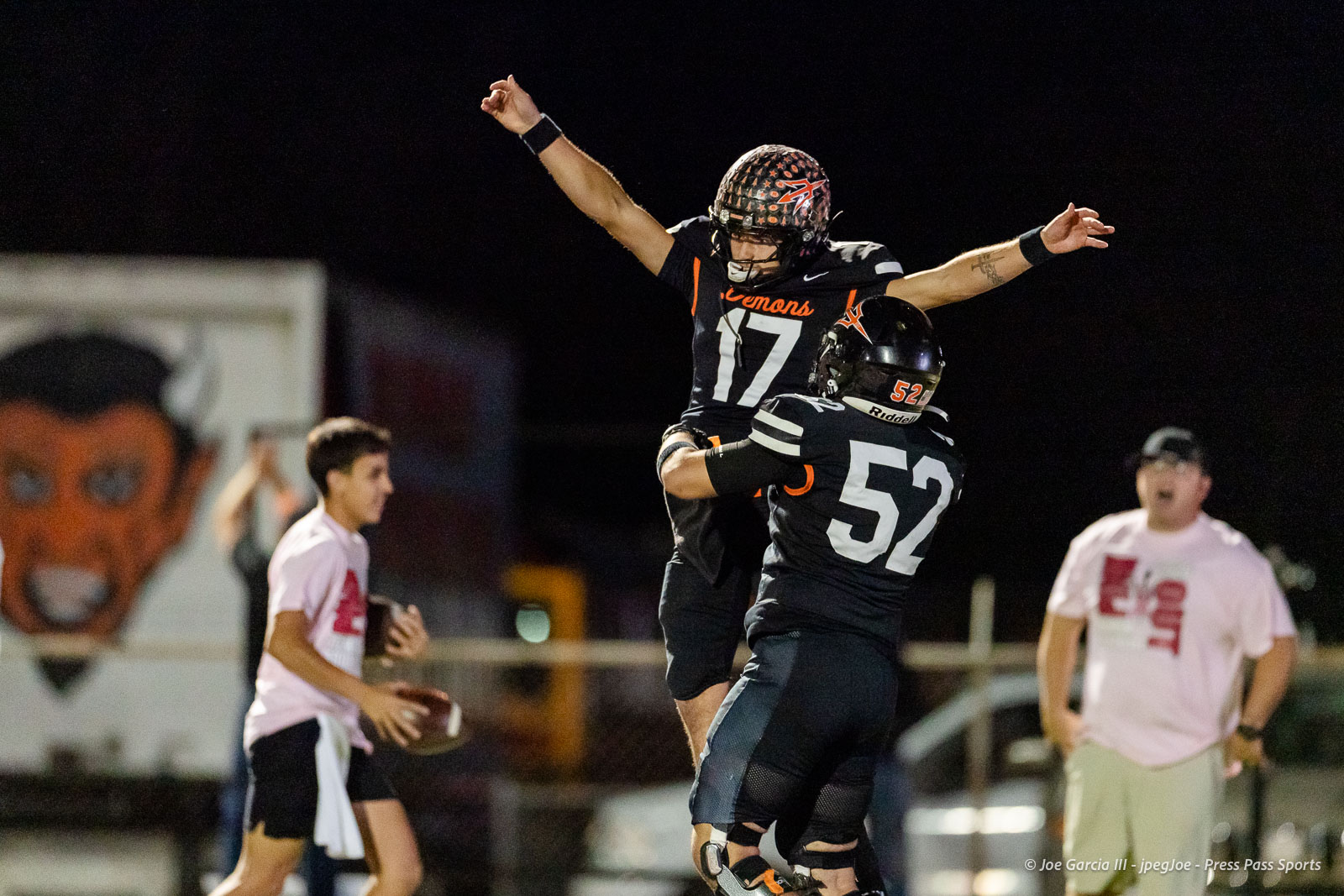 west-plains-vs-dumas-football-jpegJoe-pps-38 | Press Pass Sports Two football players in black jerseys (numbers 17 and 52) celebrate midair on a night game, arms raised in triumph.