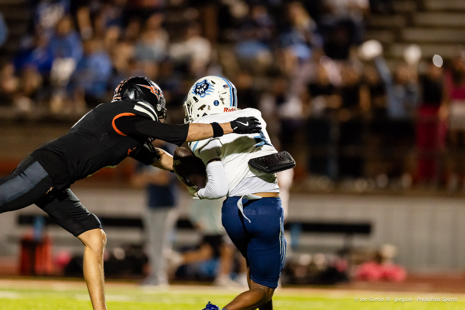 west-plains-vs-dumas-football-jpegJoe-pps-39 | Press Pass Sports Two football players collide in a tackle during a night game, one in black uniform reaching for a runner in white and blue helmets, on a crowded stadium field.