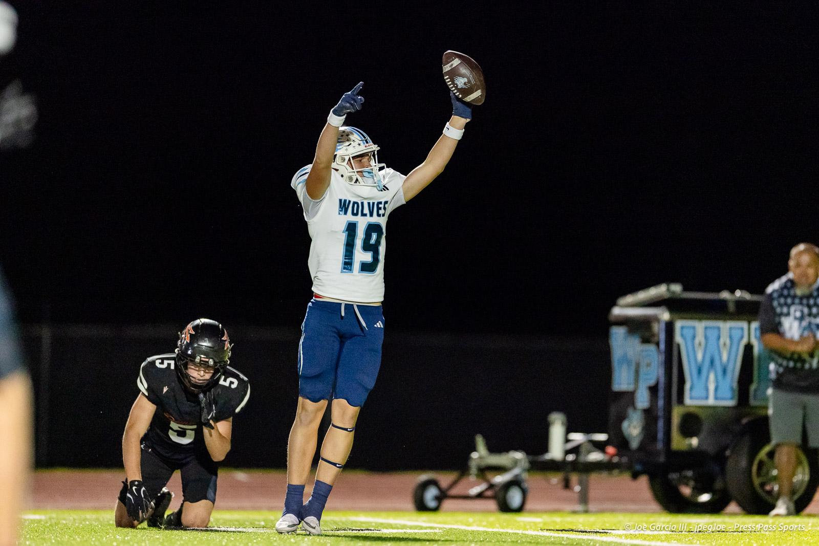 west-plains-vs-dumas-football-jpegJoe-pps-41 | Press Pass Sports Football player in a white Wolves jersey (#19) leaps with arms raised to catch a football at night, while a defender kneels nearby on the field.