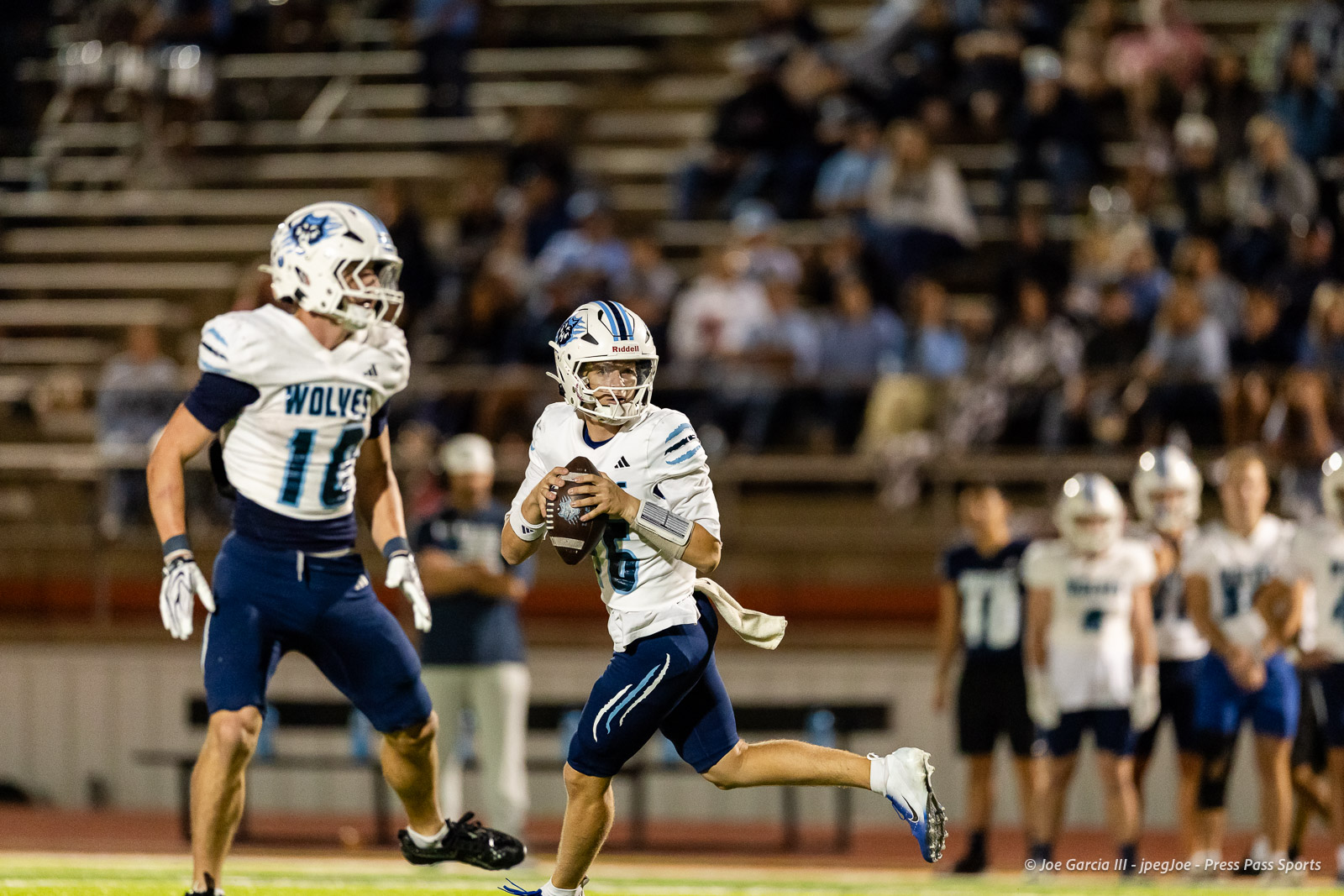 west-plains-vs-dumas-football-jpegJoe-pps-42 | Press Pass Sports Quarterback in white and blue jersey runs with the football during a game, with a teammate approaching and spectators in the stands behind.