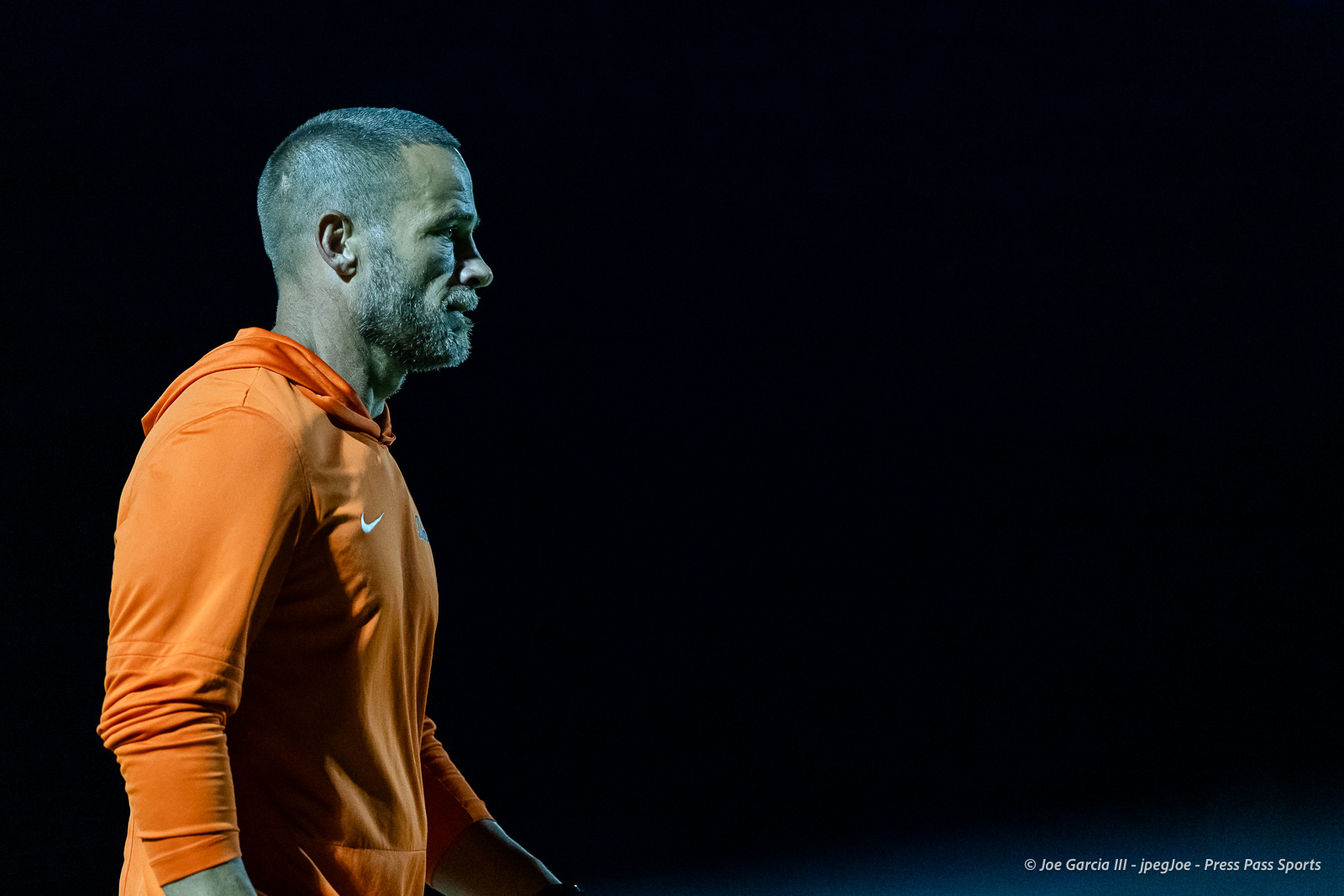 west-plains-vs-dumas-football-jpegJoe-pps-46 | Press Pass Sports Side profile of a man in an orange hoodie standing against a dark night background, illuminated by stadium lights.