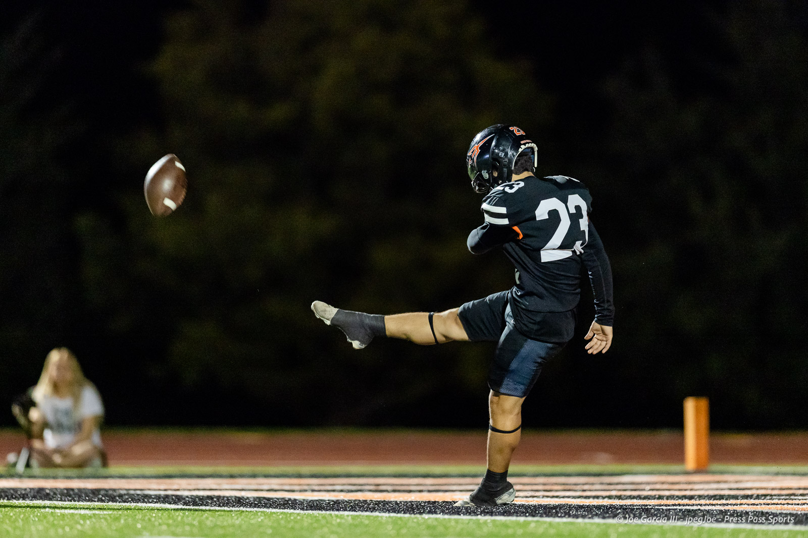 west-plains-vs-dumas-football-jpegJoe-pps-48 | Press Pass Sports Football kicker in dark uniform (#23) punts the ball during a night game on a glowing turf field, helmet on.