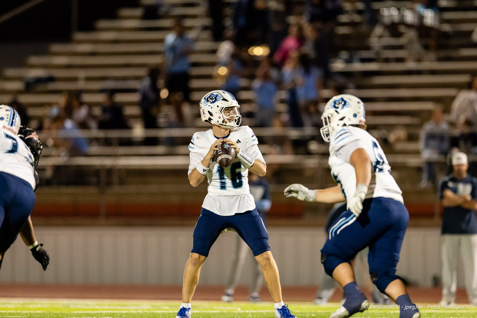 west-plains-vs-dumas-football-jpegJoe-pps-49 | Press Pass Sports Quarterback in white jersey number 16 holding a football, looking for a passer on a nighttime football field.
