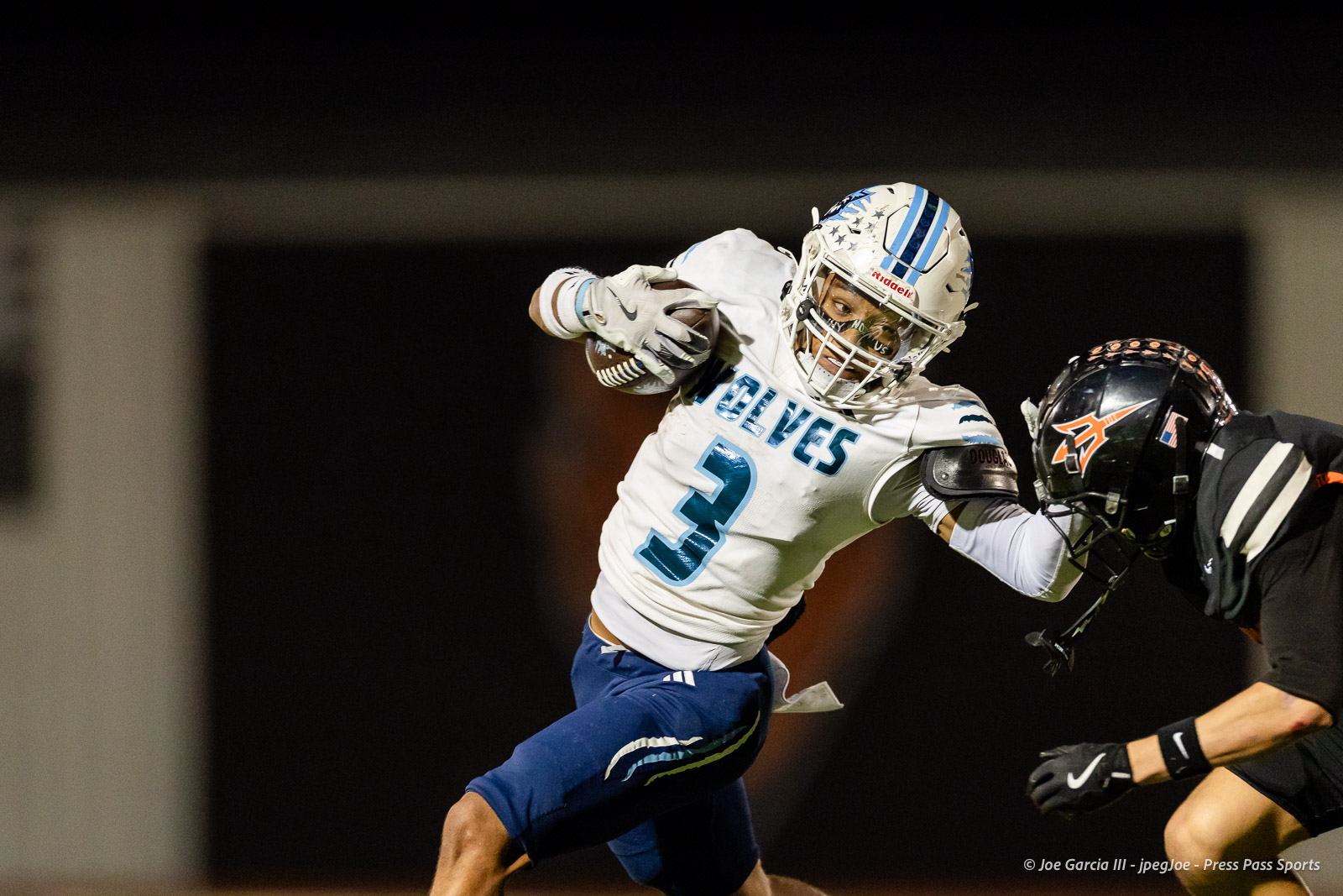 west-plains-vs-dumas-football-jpegJoe-pps-51 | Press Pass Sports A football player in a white and blue uniform with number 3 carries the ball while leaping forward, pursued by a tackler in black at night.