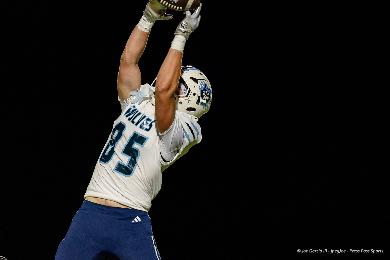 west-plains-vs-dumas-football-jpegJoe-pps-55 | Press Pass Sports Football player in white and blue, jersey number 25, leaps upward to catch a football against a dark night sky.