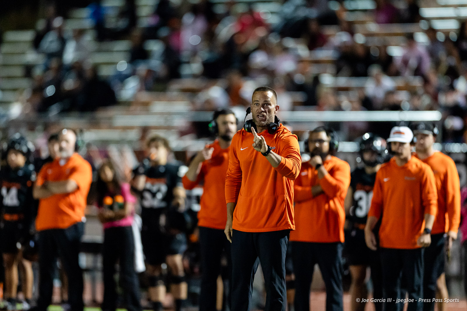 west-plains-vs-dumas-football-jpegJoe-pps-57 | Press Pass Sports Coach in an orange hoodie with a headset on, shouting and signaling from the sideline during a game.