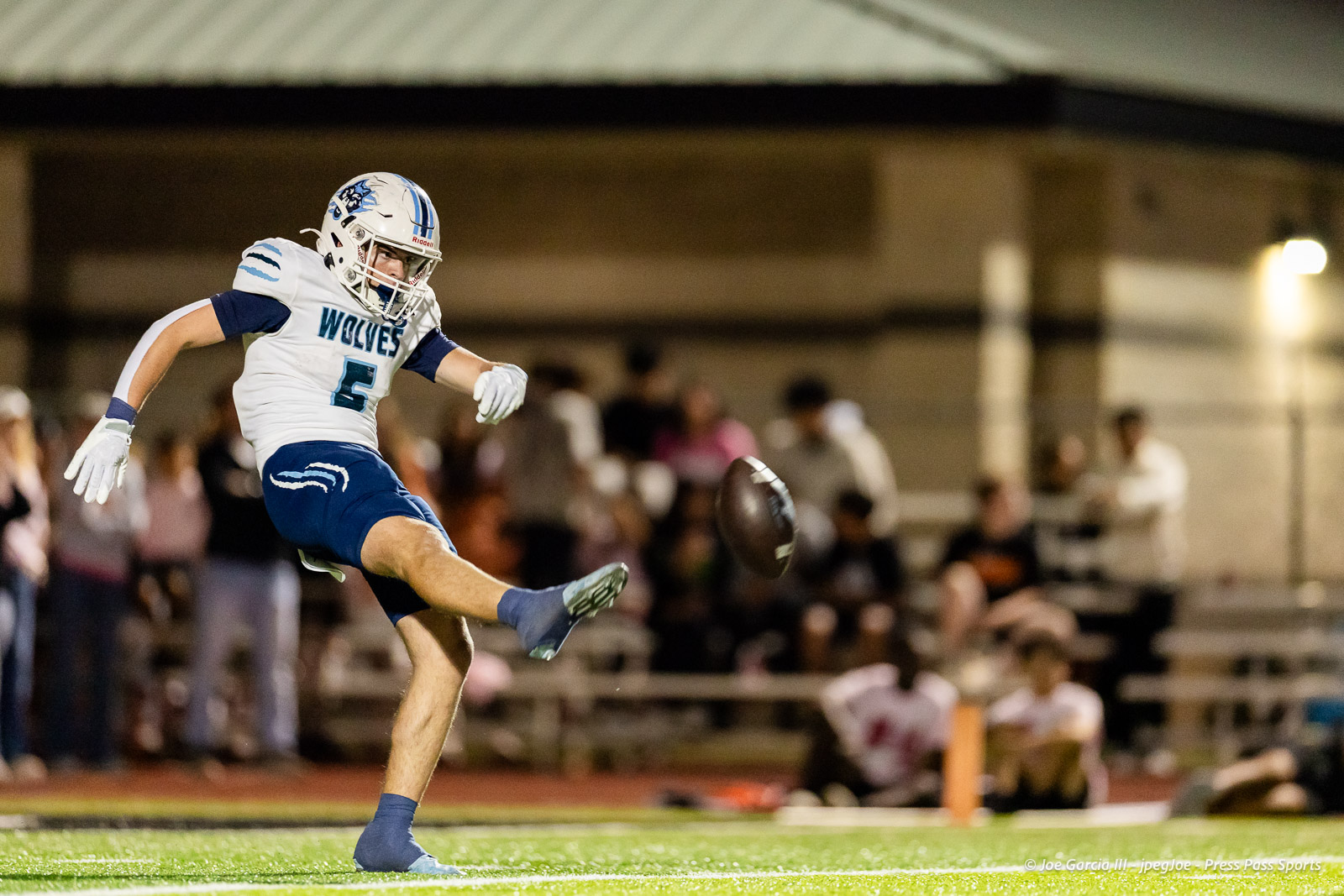 west-plains-vs-dumas-football-jpegJoe-pps-58 | Press Pass Sports American football player in a white and blue Wolves jersey (#5) kicking a football during a night game, with a blurred crowd in the background on a lit field.