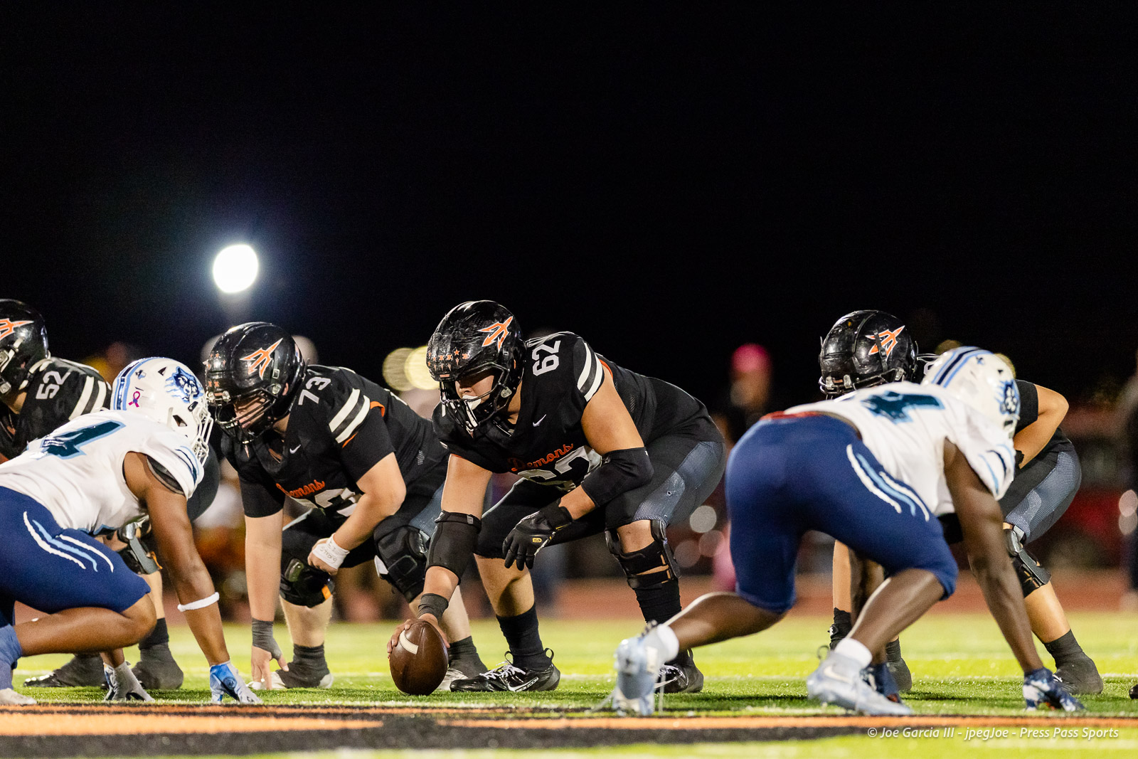 west-plains-vs-dumas-football-jpegJoe-pps-59 | Press Pass Sports Football players line up at the line of scrimmage during a night game, center poised to snap the ball behind the offensive line in black uniforms with orange accents under stadium lights.