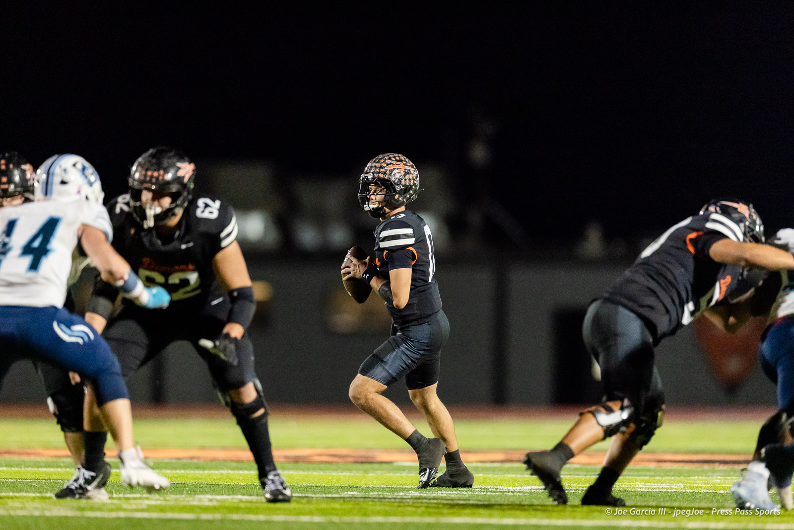 west-plains-vs-dumas-football-jpegJoe-pps-62 | Press Pass Sports Quarterback in a black uniform holding the football, looking downfield during a night game as teammates and defenders collide nearby.