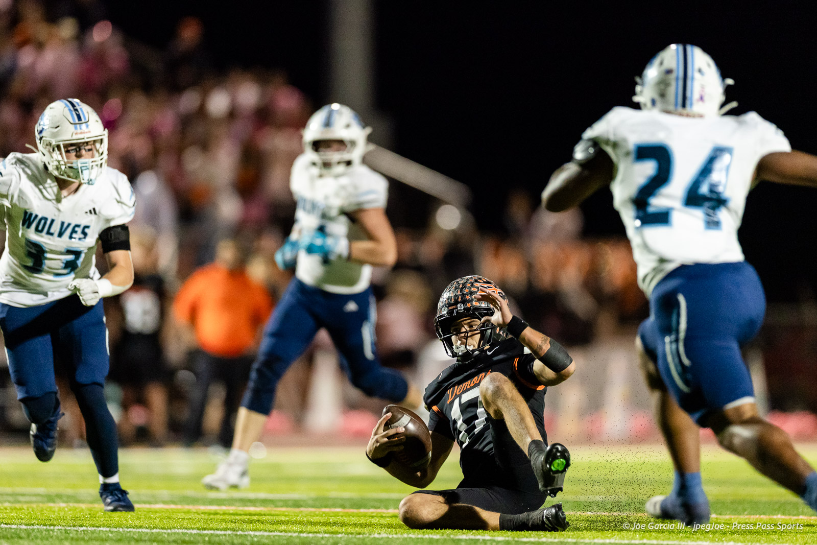 west-plains-vs-dumas-football-jpegJoe-pps-63 | Press Pass Sports Football player in black and orange sits on the turf clutching the ball during a night game, surrounded by white-uniform opponents.