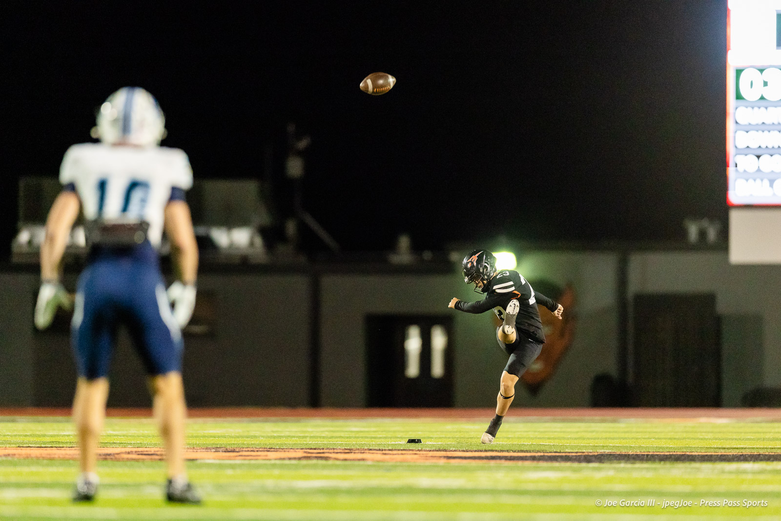 west-plains-vs-dumas-football-jpegJoe-pps-65 | Press Pass Sports A football kicker in black uniform mid-kick on a night field, ball in the air as a defender watches from in front.