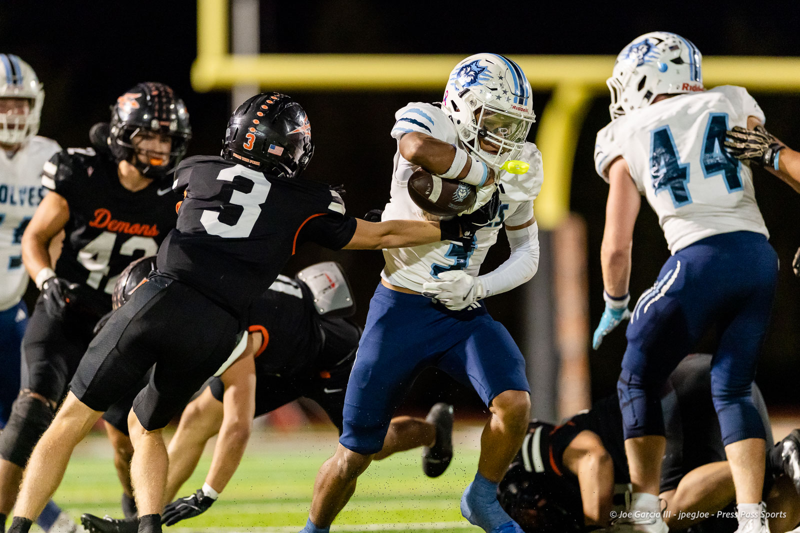 west-plains-vs-dumas-football-jpegJoe-pps-66 | Press Pass Sports Football game at night: a player in a white jersey with blue pants is tackled by a defender in a black jersey with the number 3, as others look on near the goal post backdrop.