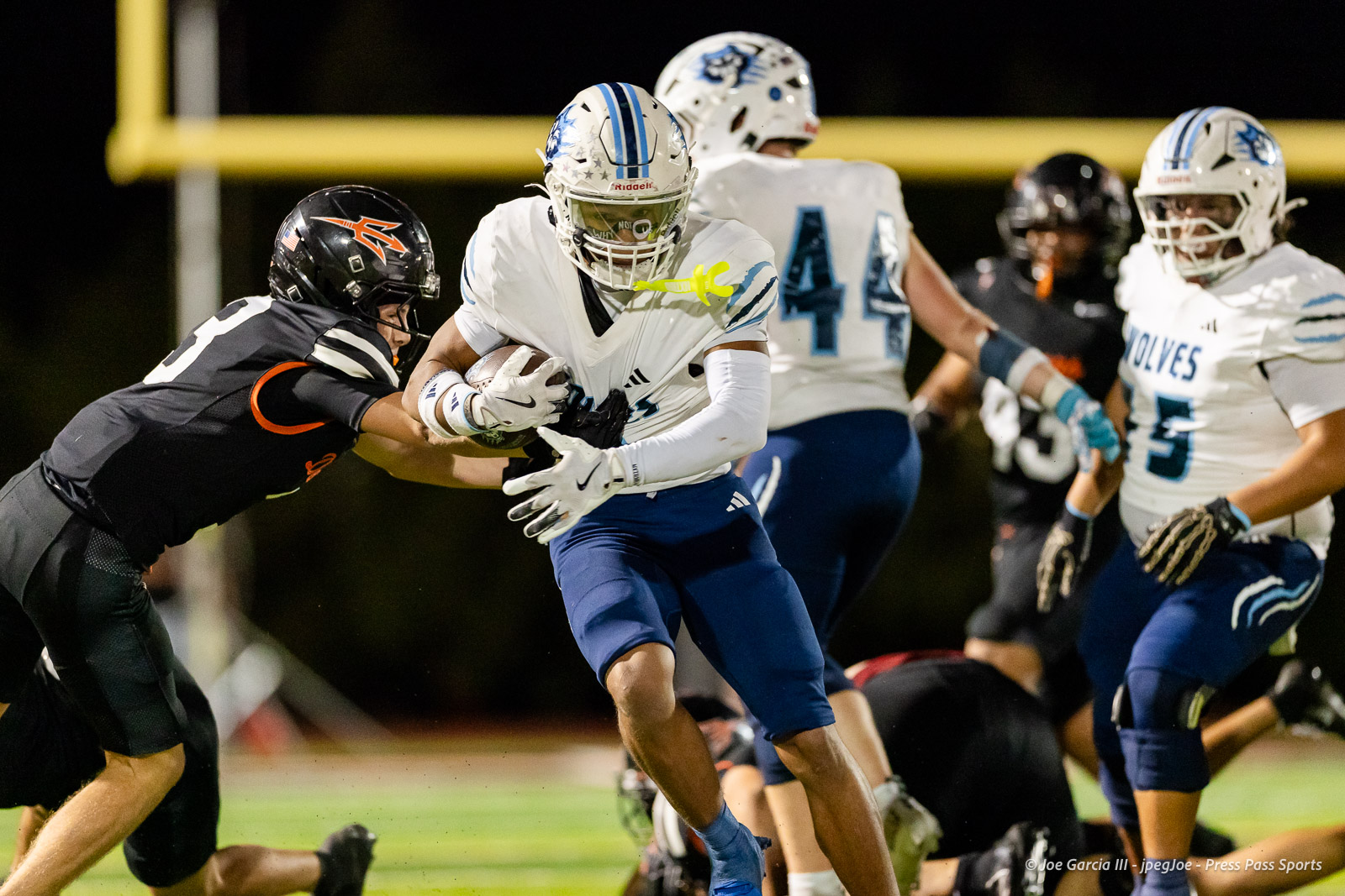 west-plains-vs-dumas-football-jpegJoe-pps-67 | Press Pass Sports Football player in white jersey and blue pants runs with the ball while being tackled by a defender in black, under stadium lights at night.