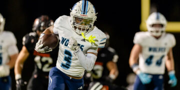 Football player in white and blue uniform (#3) carries the ball and runs under stadium lights in a night game.