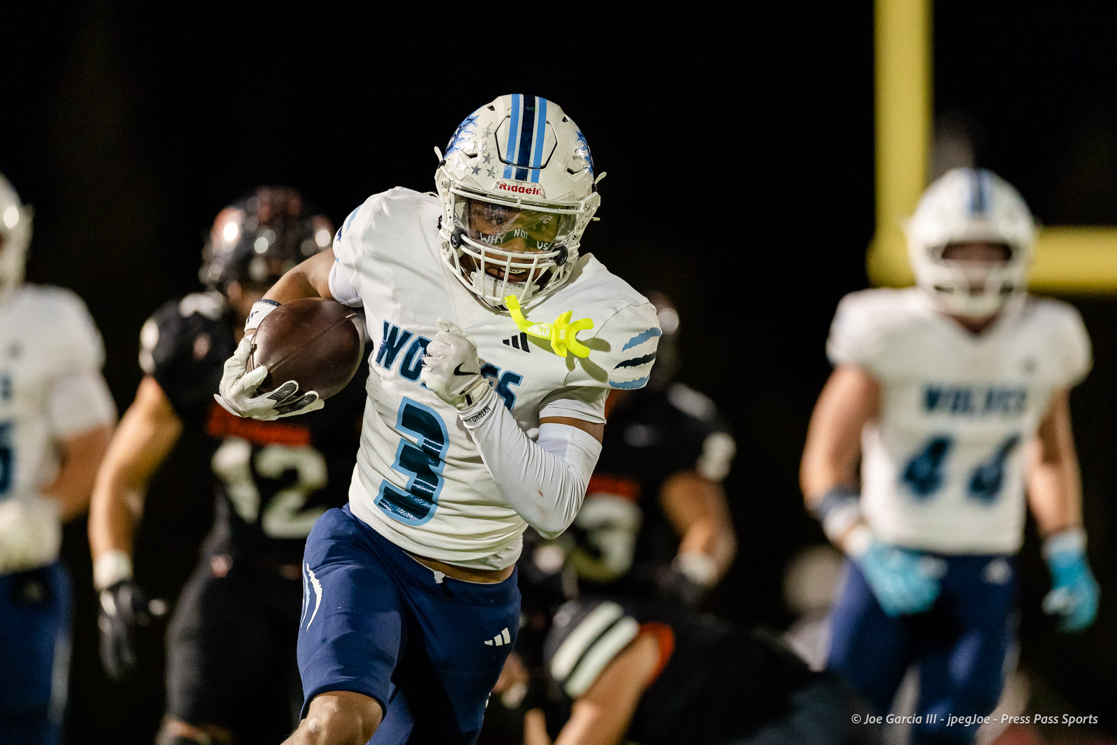 west-plains-vs-dumas-football-jpegJoe-pps-68 | Press Pass Sports Football player in white and blue uniform (#3) carries the ball and runs under stadium lights in a night game.