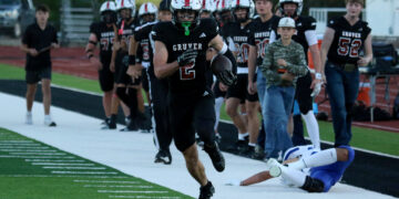 Gruver football player with number 2 runs with the ball along the sideline, teammates in pursuit behind him, on a football field.