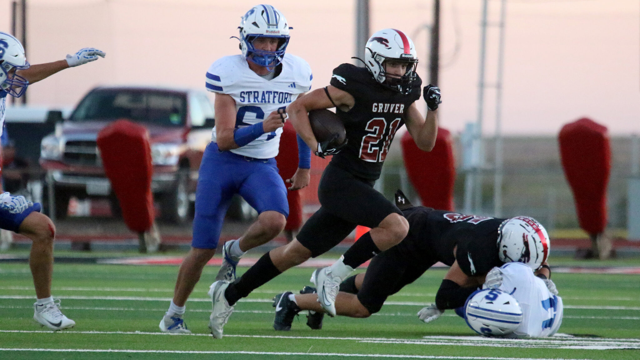 1K6A3451 | Press Pass Sports Running back in black with number 21 Gruver sprints with the ball as a Stratford defender in white and blue chases him on the turf field, with a fallen player nearby.