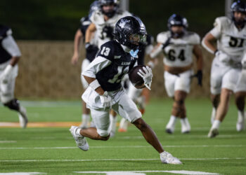 CHS-RHS-FB-43 | Press Pass Sports Football player in dark uniform carrying the ball, sprinting on a green field at night with pursuing players in white jerseys behind him.