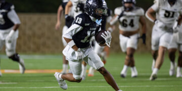 Football player in dark uniform carrying the ball, sprinting on a green field at night with pursuing players in white jerseys behind him.