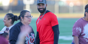 Bearded man in a red athletic shirt and cap among a group on a sports field.
