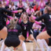 IMG_6655 | Press Pass Sports Female volleyball players in black uniforms with pink trim celebrate on a gym floor as fans clap in the stands.