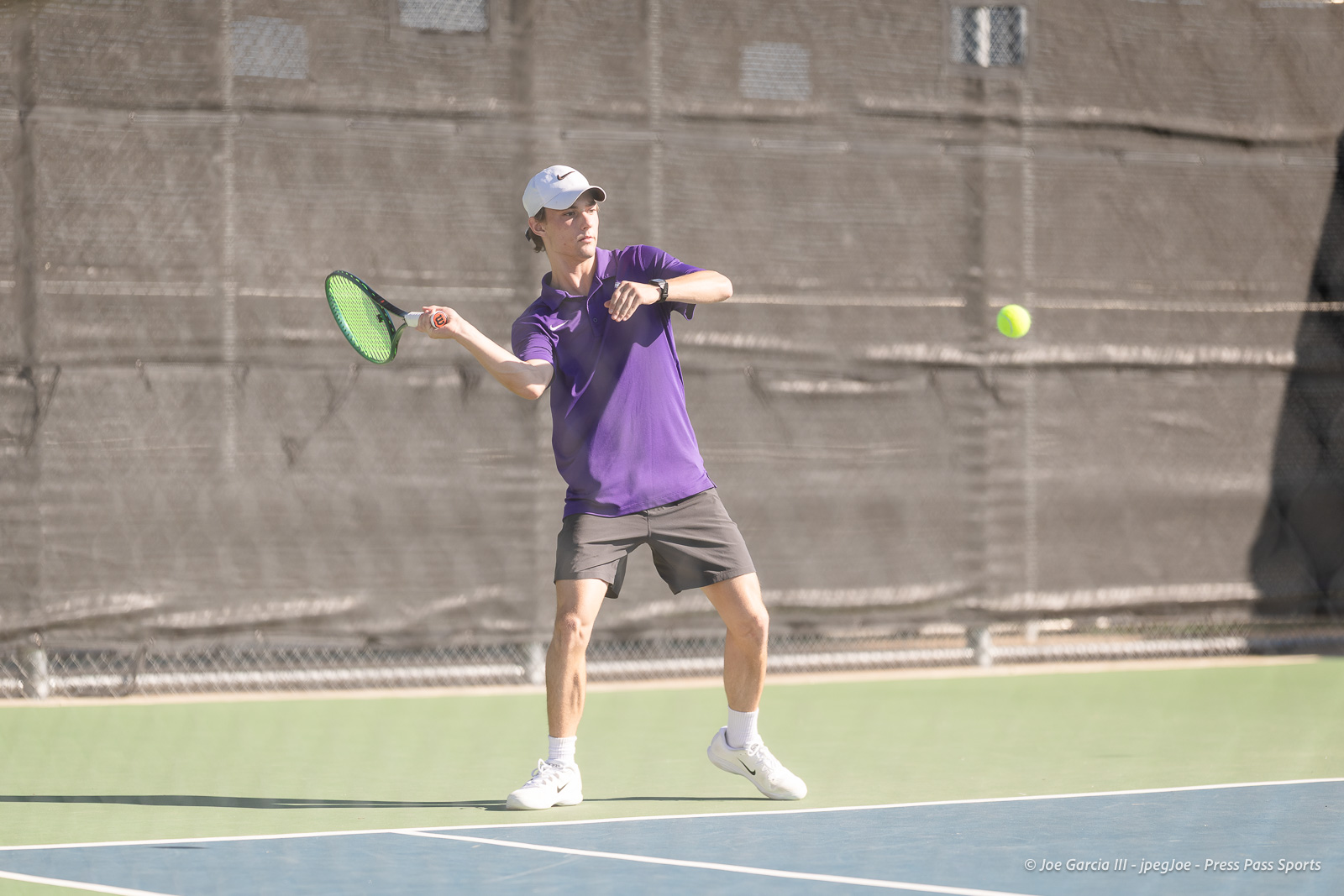 canyon-regional-tennis-1 | Press Pass Sports Tennis player in a purple polo and white cap hits a forehand on an outdoor court, green racket angled toward a bright yellow ball.