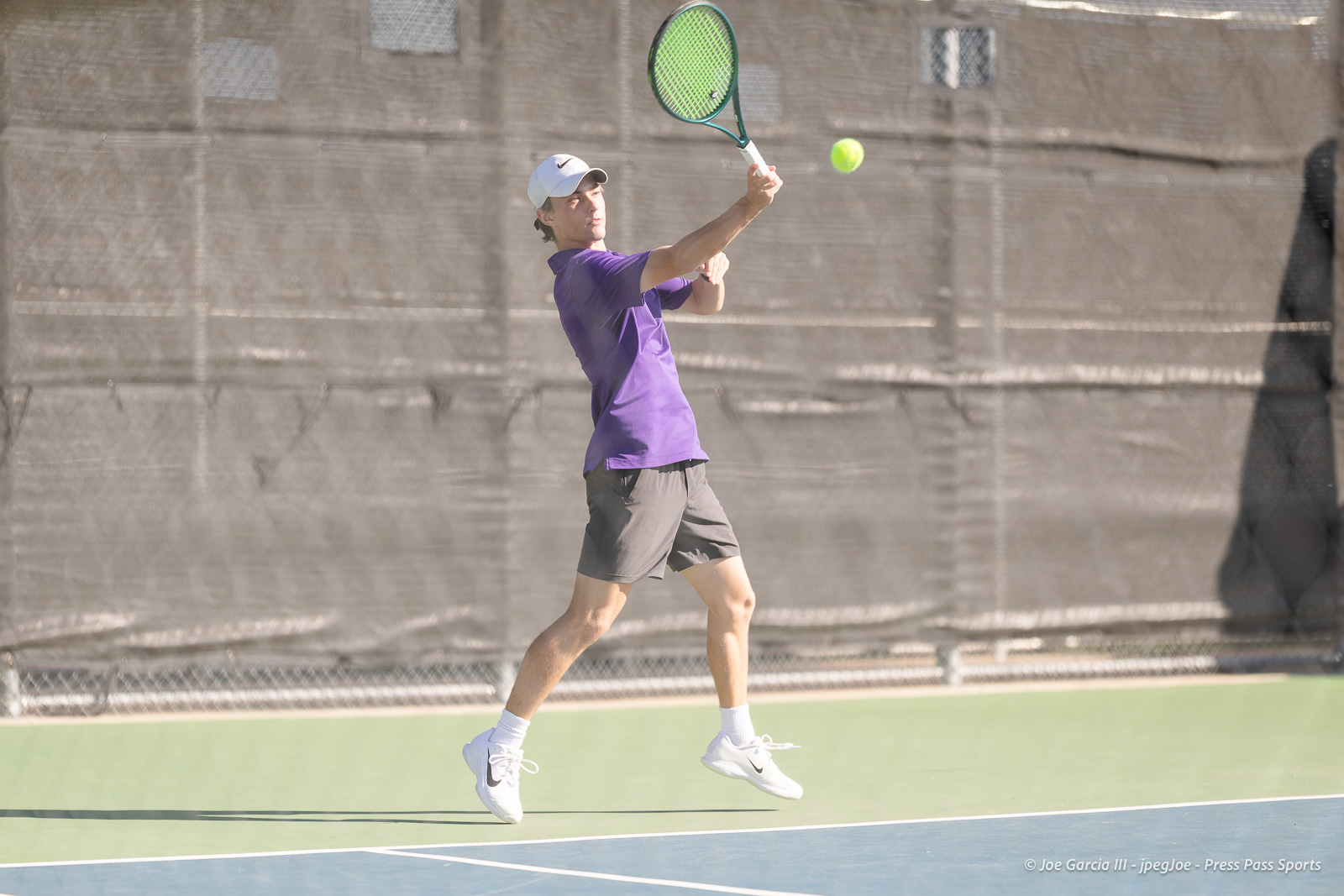 canyon-regional-tennis-2 | Press Pass Sports Tennis player in a purple shirt and gray shorts jumping to hit a forehand on an outdoor court, ball in midair near the racket.