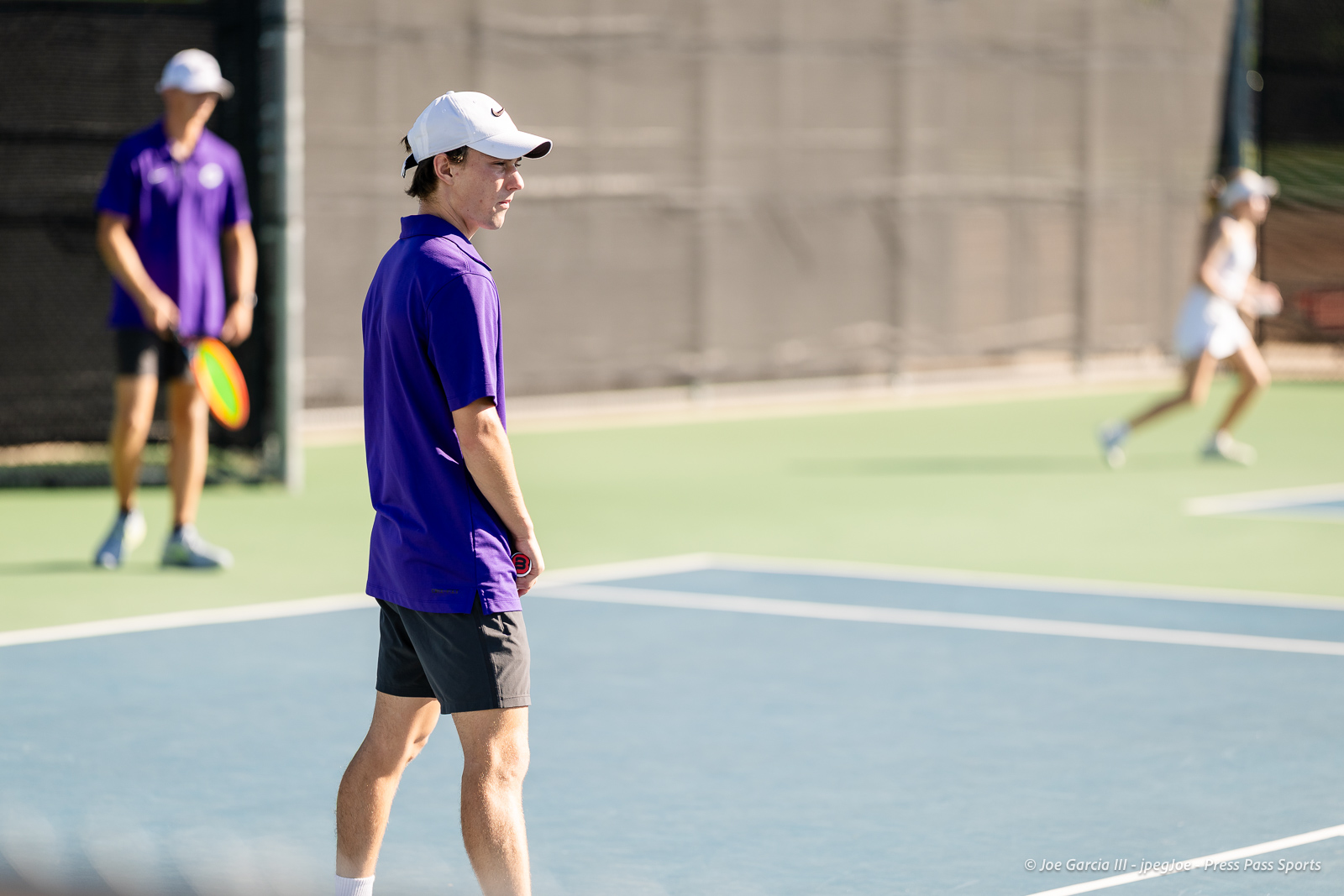 canyon-regional-tennis-3 | Press Pass Sports Man in a purple polo and white cap standing on a tennis court, facing left as players rally in the background