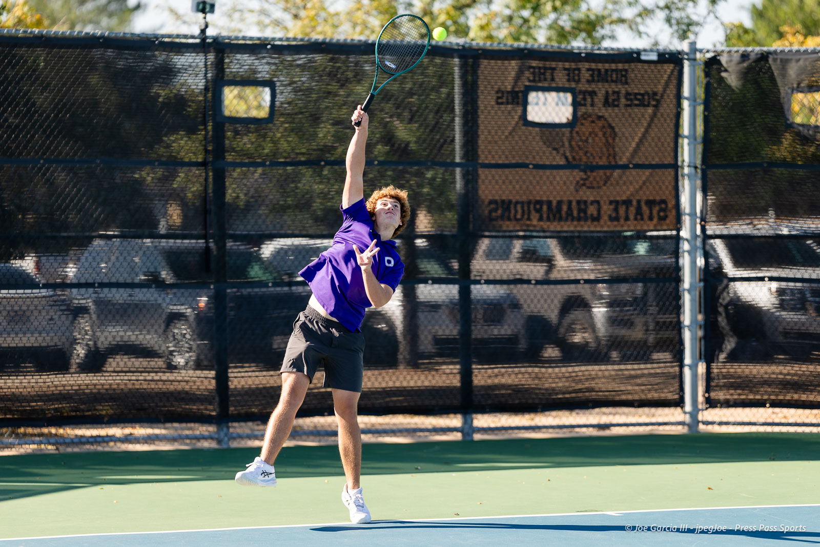 canyon-regional-tennis-4 | Press Pass Sports Young male tennis player in a purple shirt leaps to hit a ball with a racket on an outdoor court behind a chain-link fence.