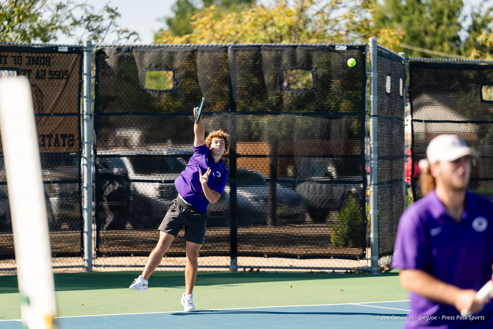 canyon-regional-tennis-6 | Press Pass Sports Young man in a purple shirt serves on a blue tennis court, ball midair against a chain-link fence behind him, sunny day.