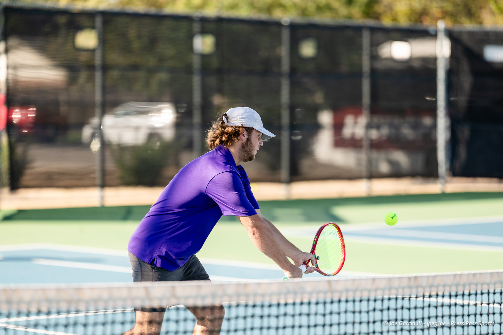 canyon-regional-tennis-7 | Press Pass Sports Tennis player in a purple shirt and white cap preparing to return a shot on a blue-green court, net in foreground, ball in midair.