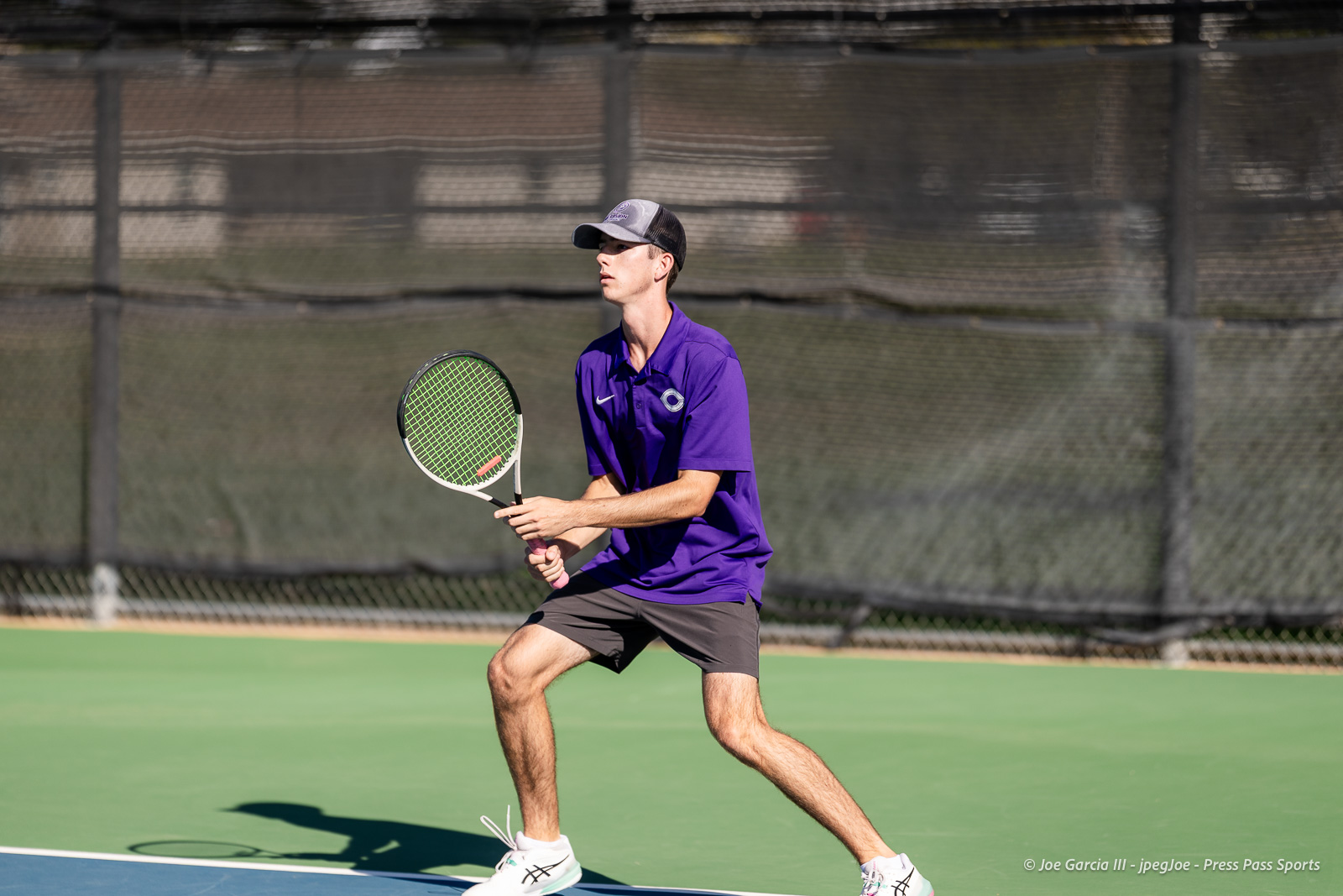 canyon-regional-tennis-8 | Press Pass Sports Male tennis player in a purple shirt ready to return a ball on an outdoor court with a green surface and black fence in background