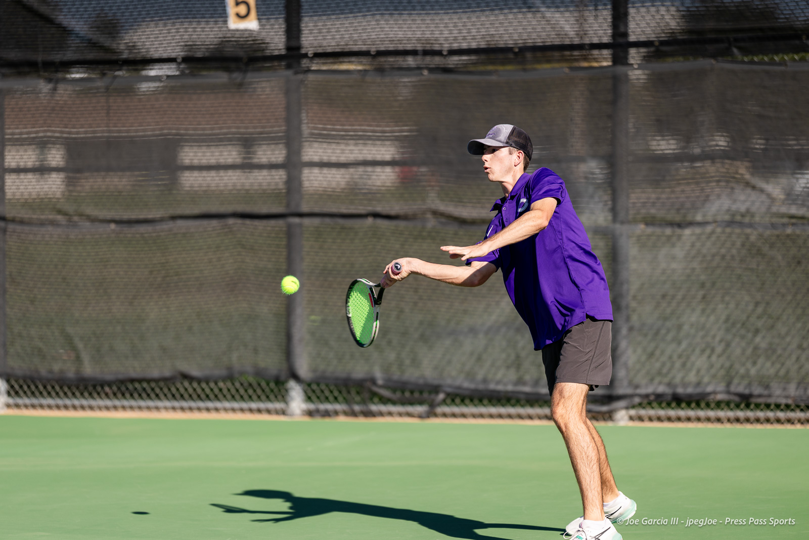 canyon-regional-tennis-10 | Press Pass Sports Male tennis player in a purple shirt and cap about to hit a forehand on an outdoor court with a bright tennis ball mid-air in front of him.