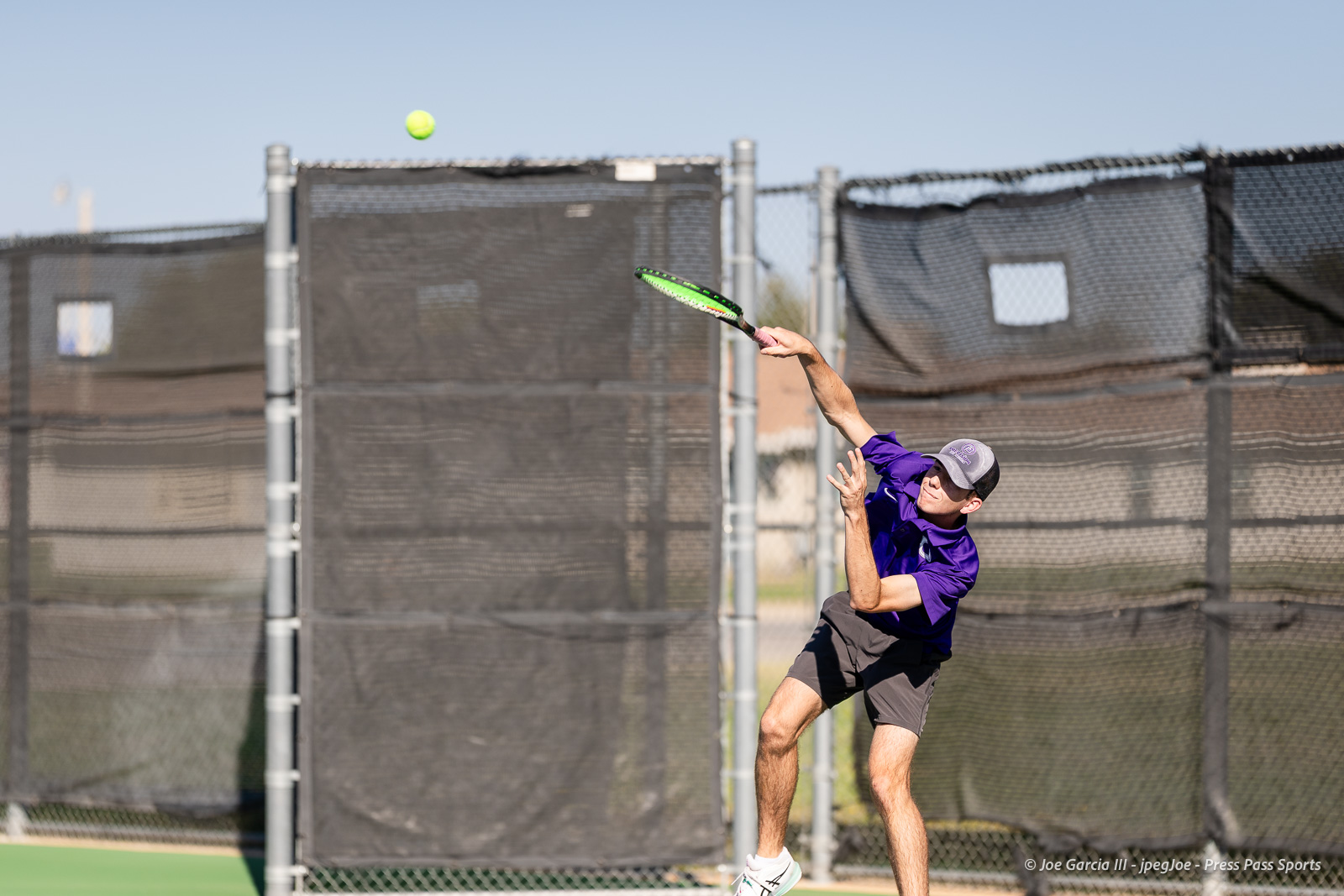canyon-regional-tennis-11 | Press Pass Sports Tennis player in a purple shirt and cap jumping to hit an overhead shot as a tennis ball flies overhead on an outdoor court behind a black chain-link fence.