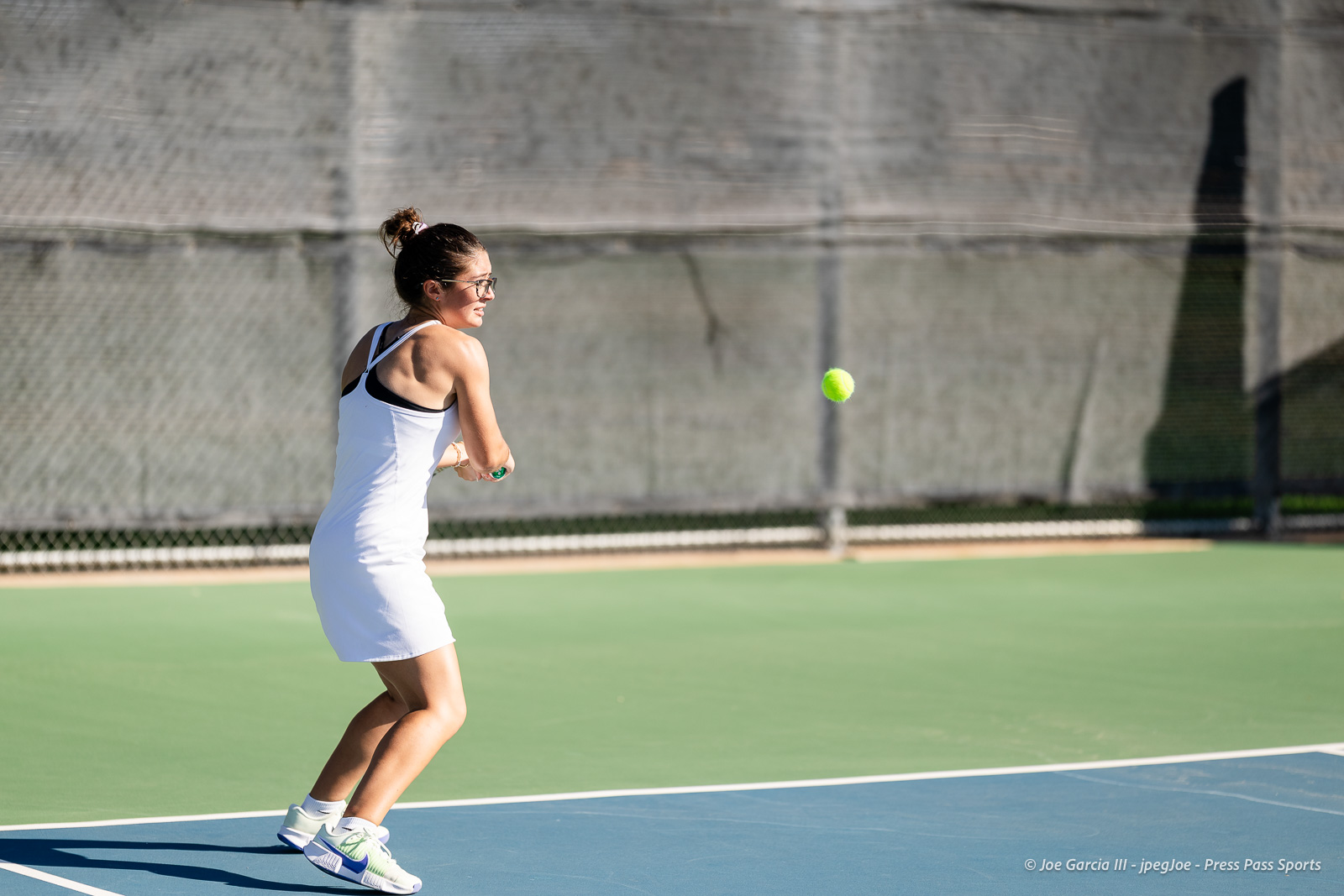 canyon-regional-tennis-12 | Press Pass Sports Woman in a white tennis dress backhands a ball on an outdoor court, neon tennis ball in midair as a fence looms in the background.