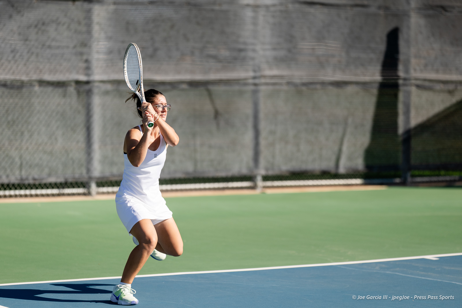 canyon-regional-tennis-14 | Press Pass Sports Female tennis player in a white outfit prepares a two-handed backhand on a blue tennis court with a chain-link fence in the background.