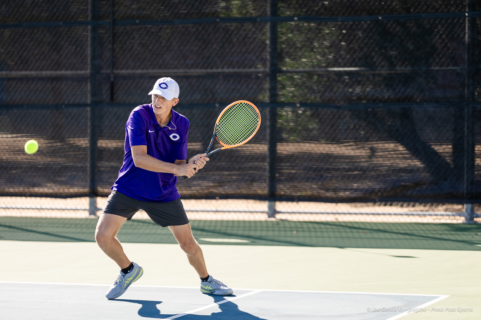 canyon-regional-tennis-16 | Press Pass Sports Tennis player in a purple shirt and white cap prepares to hit a backhand on a sunlit court with a fence backdrop behind him.