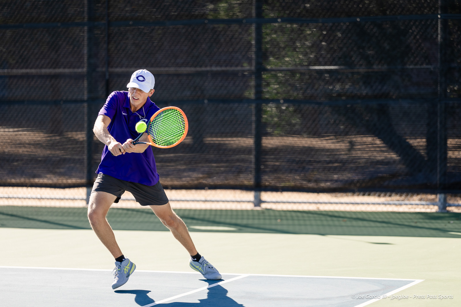 canyon-regional-tennis-17 | Press Pass Sports Male tennis player in a purple shirt and white cap lunging to return a ball on an outdoor court with a chain-link fence in the background, sunny day.