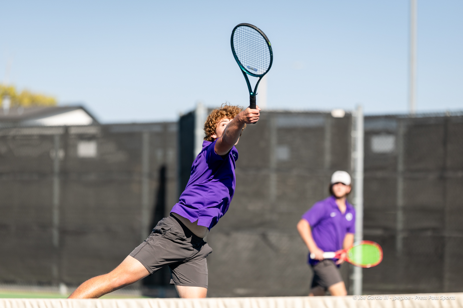 canyon-regional-tennis-18 | Press Pass Sports Tennis player in a purple shirt leaps to strike the ball with a racket on a sunny outdoor court; another player watches in the background.