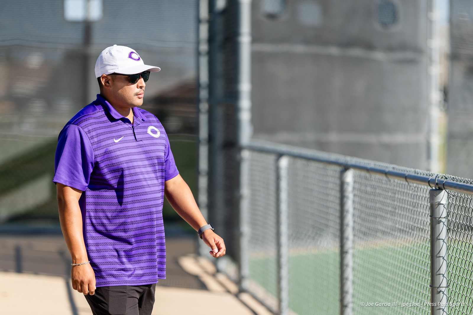 canyon-regional-tennis-19 | Press Pass Sports Man in a purple polo and white cap stands beside a chain-link fence at an outdoor sports facility, wearing sunglasses.