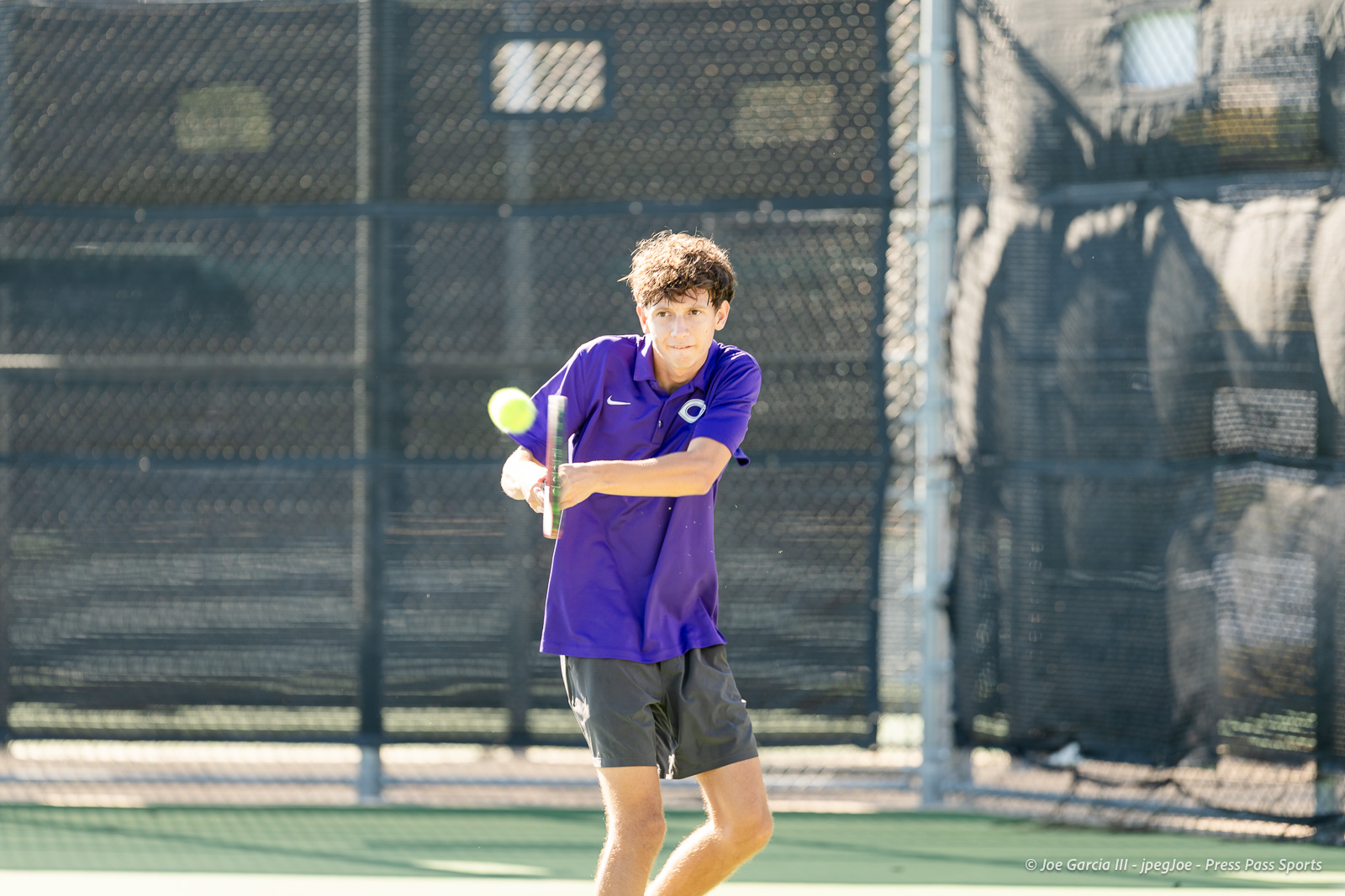 canyon-regional-tennis-22 | Press Pass Sports Young boy in a purple polo hits a tennis ball with a racket on an outdoor court, focused on the shot