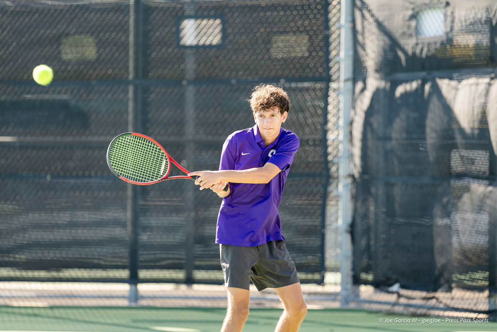 canyon-regional-tennis-23 | Press Pass Sports Teen tennis player in a purple polo swings a red-and-green racquet to return a ball on an outdoor court with a chain-link fence in the background?