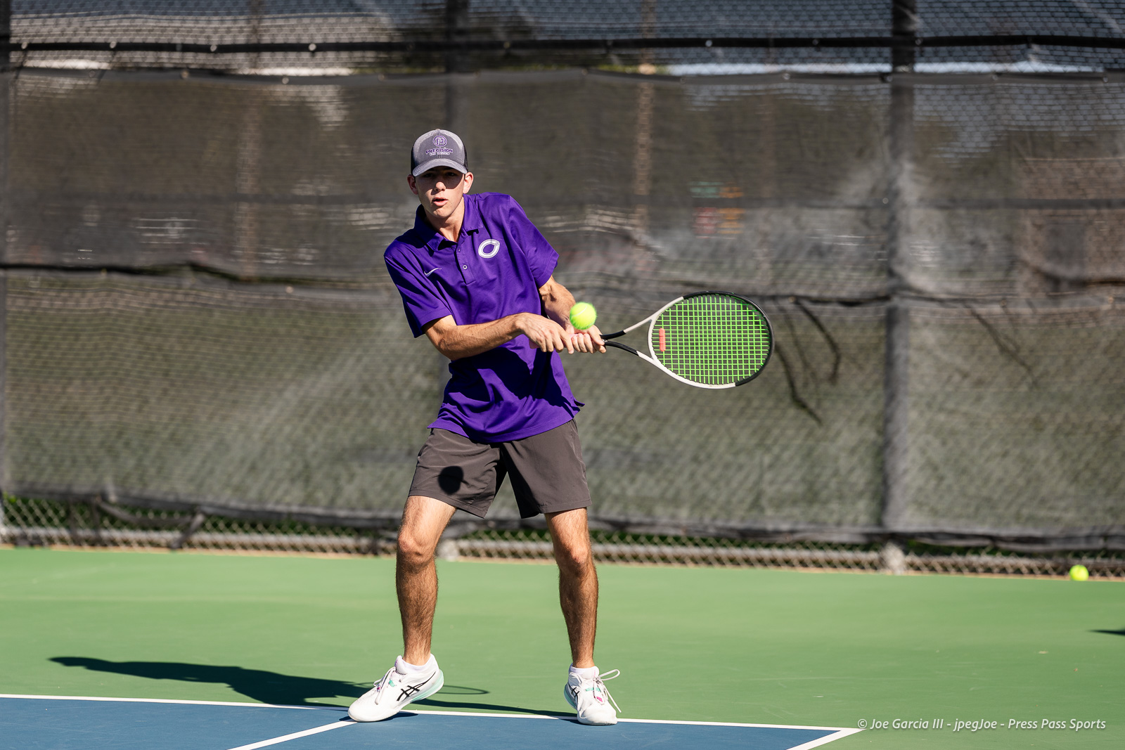 canyon-regional-tennis-24 | Press Pass Sports Male tennis player in a purple shirt hits a backhand on a green court with a chain-link fence background.
