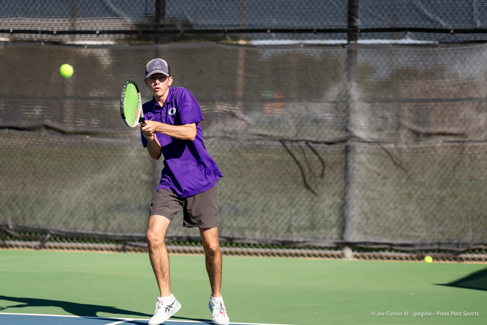canyon-regional-tennis-25 | Press Pass Sports Male tennis player in a purple polo and gray cap prepares to volley on an outdoor court as a bright ball approaches left of him.